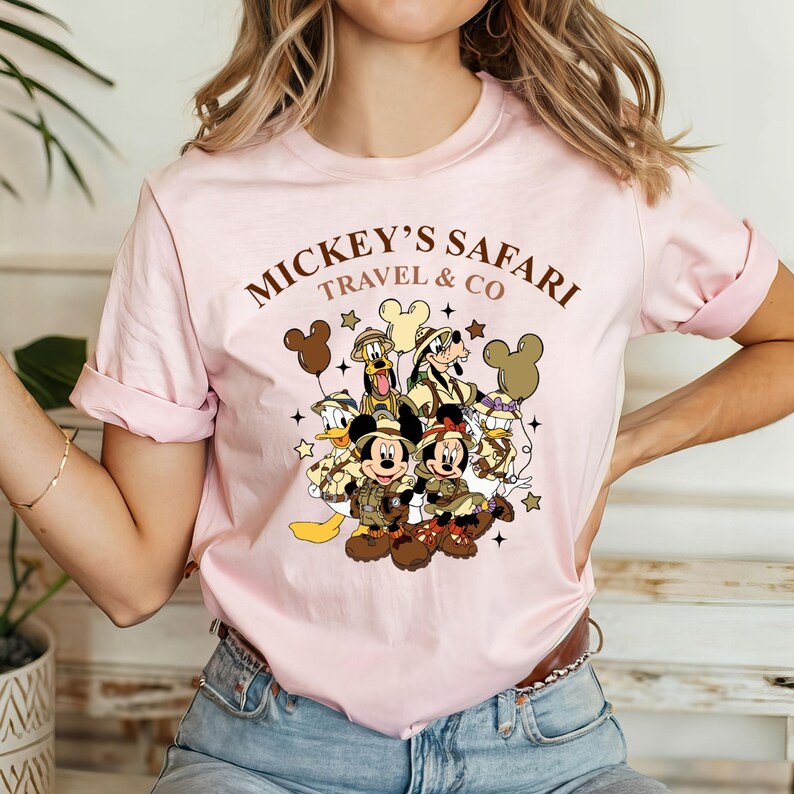 K&ouml;nnte beinhalten: Rosa T-Shirt mit einem Aufdruck von Mickey Mouse und seinen Freunden in Safari-Outfits. Der Text "Mickey's Safari Travel & Co" ist auf dem Shirt aufgedruckt.