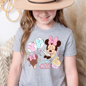 May include: A grey t-shirt with a pink and blue Minnie Mouse design. The design includes Minnie Mouse holding balloons, a Mickey Mouse ice cream cone, and a ticket that says "Magic".