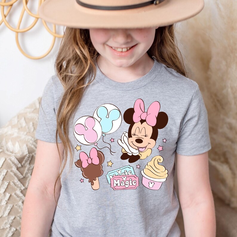 May include: A grey t-shirt with a pink and blue Minnie Mouse design. The design includes Minnie Mouse holding balloons, a Mickey Mouse ice cream cone, and a ticket that says "Magic".