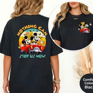 Pode incluir: T-shirt preto com um gr&aacute;fico colorido de Mickey e Minnie Mouse num carro vermelho, com o texto "NOTHING CAN STOP US NOW!". O design inclui um fundo de p&ocirc;r do sol amarelo e laranja. A t-shirt &eacute; da marca Comfort Colors.