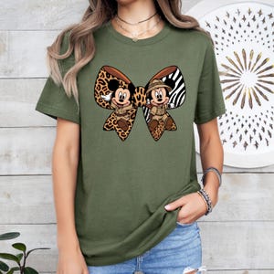 May include: Olive green t-shirt with a large bow design featuring Mickey and Minnie Mouse in safari attire. The bow has a leopard print and zebra stripe pattern. The shirt is a short-sleeved crew neck.