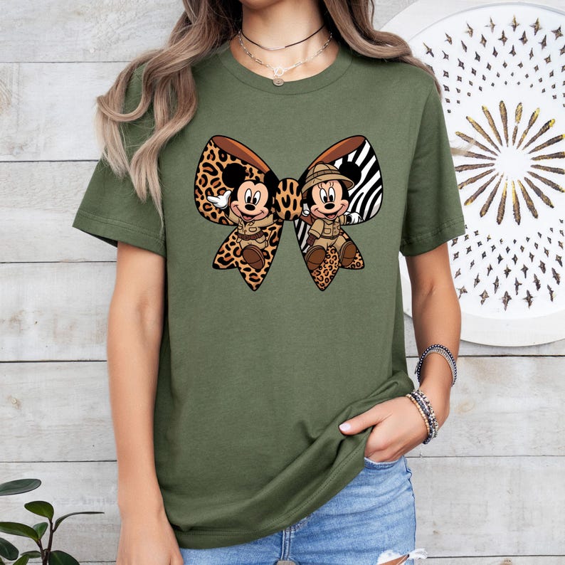 May include: Olive green t-shirt with a large bow design featuring Mickey and Minnie Mouse in safari attire. The bow has a leopard print and zebra stripe pattern. The shirt is a short-sleeved crew neck.
