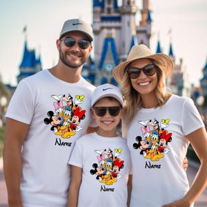 Op de afbeelding: Een gezin van drie personen draagt witte T-shirts met een kleurrijk cartoonbeeld van Mickey Mouse, Minnie Mouse, Donald Duck, Goofy en Pluto. De shirts hebben het woord "Naam" erop gedrukt.