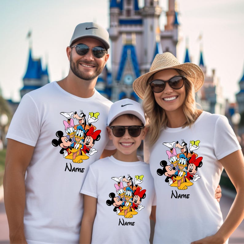 Op de afbeelding: Een gezin van drie personen draagt witte T-shirts met een kleurrijk cartoonbeeld van Mickey Mouse, Minnie Mouse, Donald Duck, Goofy en Pluto. De shirts hebben het woord "Naam" erop gedrukt.