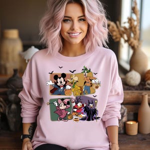 K&ouml;nnte beinhalten: Rosafarbener Pullover mit einem Grafikdruck von Mickey Mouse, Pluto, Goofy, Donald Duck und Maleficent in Halloween-Kost&uuml;men. Die Figuren halten K&uuml;rbisse und S&uuml;&szlig;igkeiten.