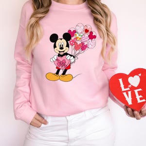 May include: Pink sweatshirt with a Mickey Mouse graphic holding a heart-shaped box of chocolates and a bunch of heart-shaped balloons. The sweatshirt has a crew neck and long sleeves.