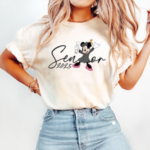 May include: White t-shirt featuring a black and white illustration of Mickey Mouse wearing a graduation cap and gown. The text "Senior 2023" is written in black cursive.