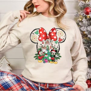 Puede incluir: Una sudadera blanca con un dise&ntilde;o navide&ntilde;o que presenta a Mickey Mouse y Minnie Mouse frente al Castillo de Cenicienta. El dise&ntilde;o incluye un lazo rojo con lunares blancos, un &aacute;rbol de Navidad y regalos.