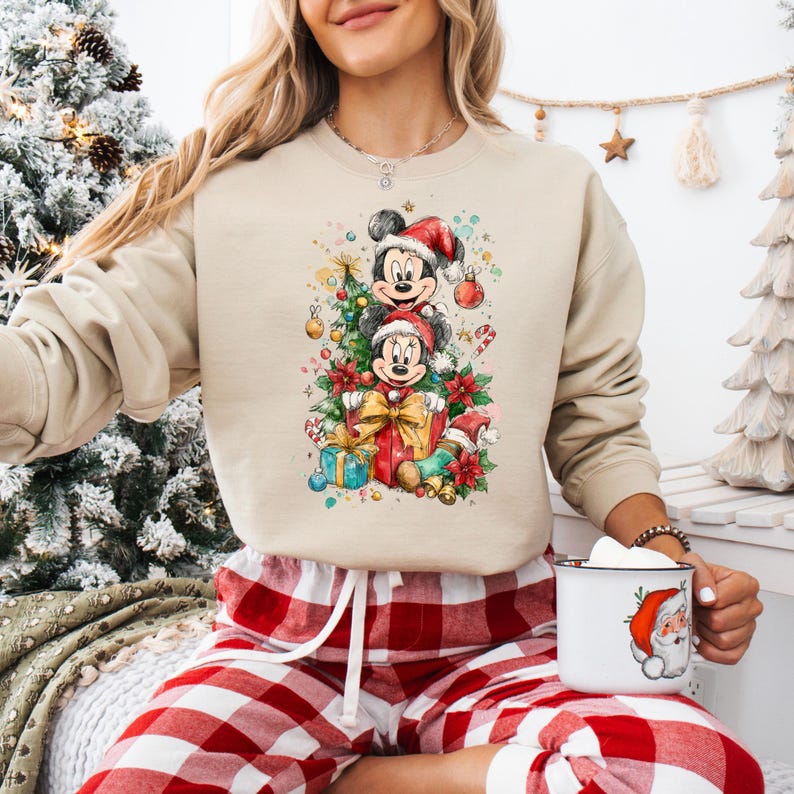 May include: Beige sweatshirt featuring a festive watercolor design of Mickey and Minnie Mouse in Santa hats, surrounded by Christmas decorations. The design includes a Christmas tree, gifts, and ornaments. The person is wearing red and white plaid pajama pants and holding a mug with a Santa Claus design.