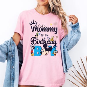 May include: Pink t-shirt with a graphic of Mickey Mouse and other Disney characters. The text on the shirt reads "Mommy of the Birthday Boy".