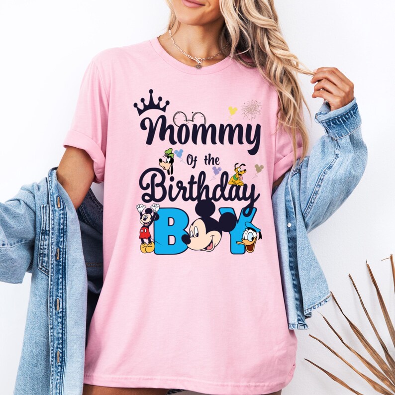 May include: Pink t-shirt with a graphic of Mickey Mouse and other Disney characters. The text on the shirt reads "Mommy of the Birthday Boy".