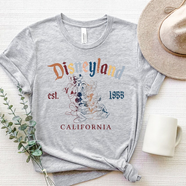May include: Gray t-shirt with a vintage-style graphic of Mickey Mouse and friends in front of the Disneyland logo. The text reads "Disneyland est. 1955 California".