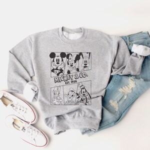 K&ouml;nnte beinhalten: Ein graues Sweatshirt mit einem schwarz-wei&szlig;en Grafikdruck von Mickey Mouse, Minnie Mouse, Donald Duck und Goofy. Die Grafik enth&auml;lt den Text "Mickey & Co. Est. 1928".