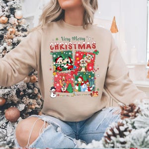 May include: Beige sweatshirt featuring a festive design with Disney characters. The graphic includes Mickey and Minnie Mouse, Chip and Dale, Pluto, Donald Duck, and more, with the text "Very Merry Christmas" in a decorative font. The design is set against a backdrop of Christmas trees and snowflakes.