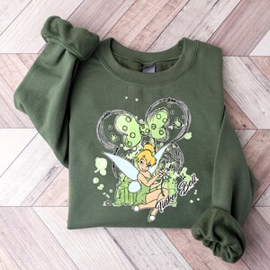 May include: Green sweatshirt with a cartoon image of Tinker Bell holding a flower. The image is surrounded by a black and white design with the words "Tinker Bell" written below.