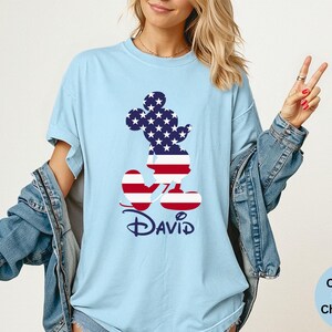 May include: Light blue t-shirt featuring a Mickey Mouse silhouette filled with a stars and stripes design. The name "David" is printed in navy blue script below the character. The shirt is a Comfort Colors Chambray.
