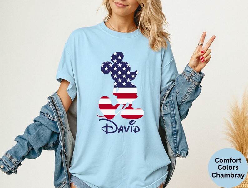 May include: Light blue t-shirt featuring a Mickey Mouse silhouette filled with a stars and stripes design. The name "David" is printed in navy blue script below the character. The shirt is a Comfort Colors Chambray.