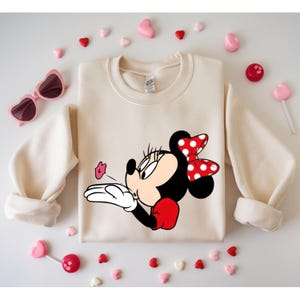 May include: Cream-colored sweatshirt featuring a Minnie Mouse graphic blowing a kiss. Minnie Mouse has a red and white polka dot bow, red gloves, and a black face. Pink heart-shaped sunglasses and candies surround the sweatshirt.