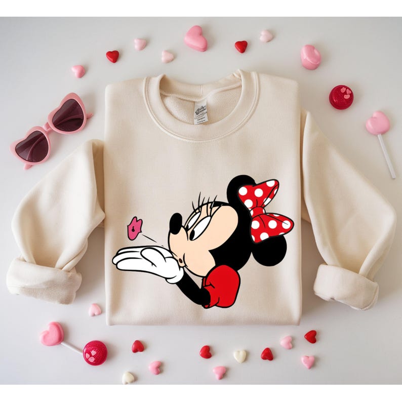 May include: Cream-colored sweatshirt featuring a Minnie Mouse graphic blowing a kiss. Minnie Mouse has a red and white polka dot bow, red gloves, and a black face. Pink heart-shaped sunglasses and candies surround the sweatshirt.