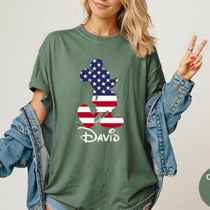 May include: Moss green t-shirt featuring a Mickey Mouse silhouette filled with a US flag design. The name "David" is printed in white script below the graphic. The shirt is part of the Comfort Colors collection.