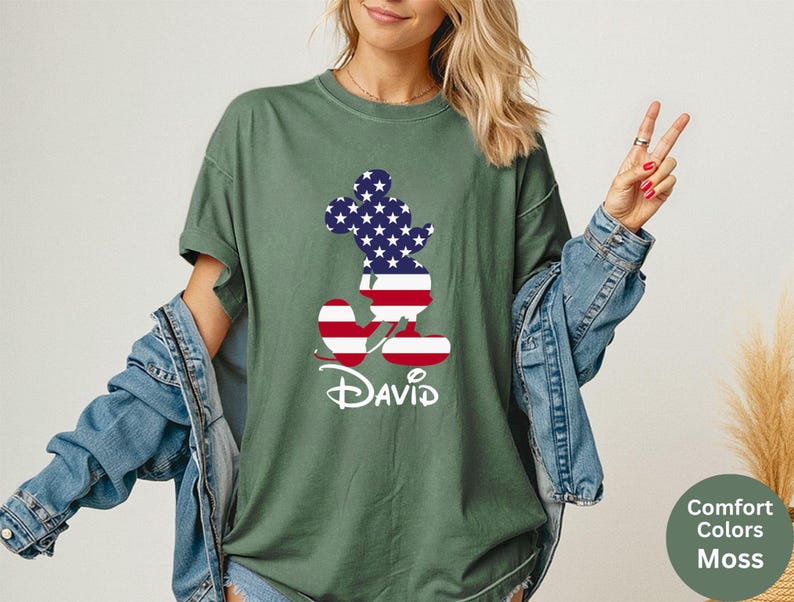 May include: Moss green t-shirt featuring a Mickey Mouse silhouette filled with a US flag design. The name "David" is printed in white script below the graphic. The shirt is part of the Comfort Colors collection.