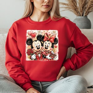 May include: Red sweatshirt featuring a graphic of Mickey and Minnie Mouse in a romantic pose. Mickey holds roses, and Minnie holds a box of chocolates. The design includes hearts and balloons, perfect for Valentine's Day or Disney fans.