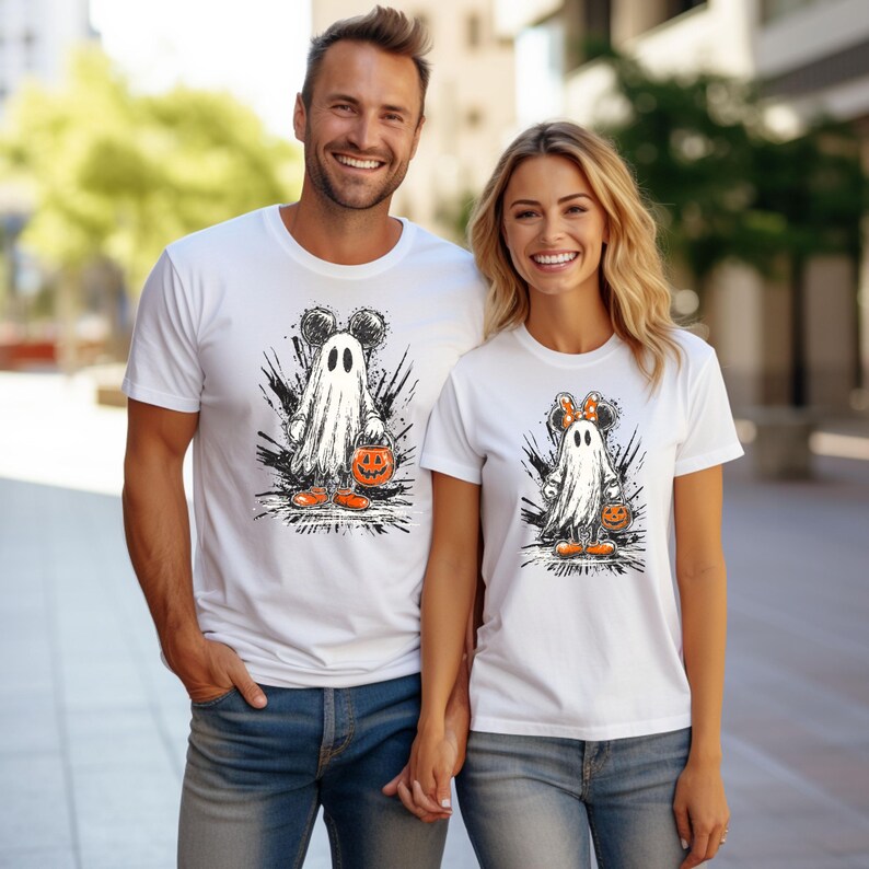Peut inclure: T-shirts blancs avec un motif d'Halloween. Le motif repr&eacute;sente un fant&ocirc;me avec des oreilles de Mickey Mouse tenant une citrouille. Le fant&ocirc;me est entour&eacute; de coups de pinceau noirs. Le t-shirt de la femme a un n&oelig;ud Minnie Mouse.