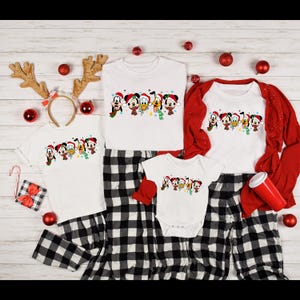 May include: A set of four white t-shirts with a Christmas-themed design featuring Mickey Mouse, Donald Duck, Goofy, and Pluto wearing Santa hats. The shirts are arranged in a family size order, with the largest shirt on top and the smallest shirt on the bottom. The shirts are all on a white wooden background with red and white plaid pants and a red mug.