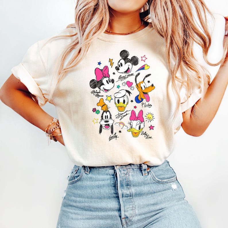 May include: A white t-shirt with a colorful cartoon graphic of Mickey Mouse, Minnie Mouse, Donald Duck, Goofy, and Pluto. The graphic includes hearts, stars, and rainbows. The text "Mickey Mouse", "Minnie Mouse", "Donald Duck", "Goofy", and "Pluto" is written in a handwritten style.