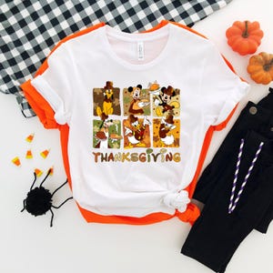 May include: White t-shirt with a Thanksgiving graphic featuring cartoon characters. The design includes six squares with characters like Mickey Mouse and Donald Duck, and the word "THANKSGIVING" in brown letters. The shirt is paired with black pants and decorative pumpkins.