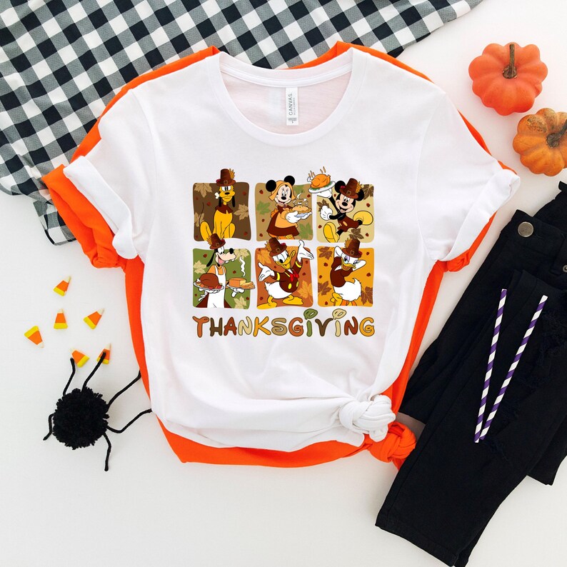 May include: White t-shirt with a Thanksgiving graphic featuring cartoon characters. The design includes six squares with characters like Mickey Mouse and Donald Duck, and the word "THANKSGIVING" in brown letters. The shirt is paired with black pants and decorative pumpkins.