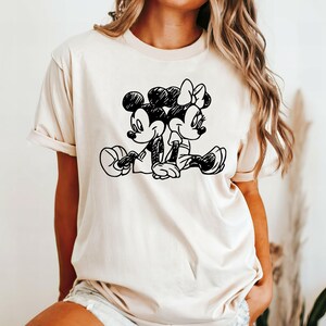 May include: A cream colored t-shirt with a black and white illustration of Mickey Mouse and Minnie Mouse holding hands.