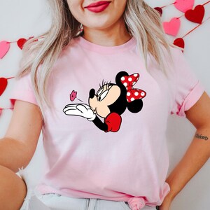 May include: A light pink t-shirt featuring a Minnie Mouse graphic. Minnie is depicted blowing a kiss, with a red and white polka dot bow, red gloves, and a black body. The graphic is centered on the shirt.