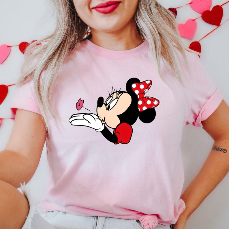 May include: A light pink t-shirt featuring a Minnie Mouse graphic. Minnie is depicted blowing a kiss, with a red and white polka dot bow, red gloves, and a black body. The graphic is centered on the shirt.