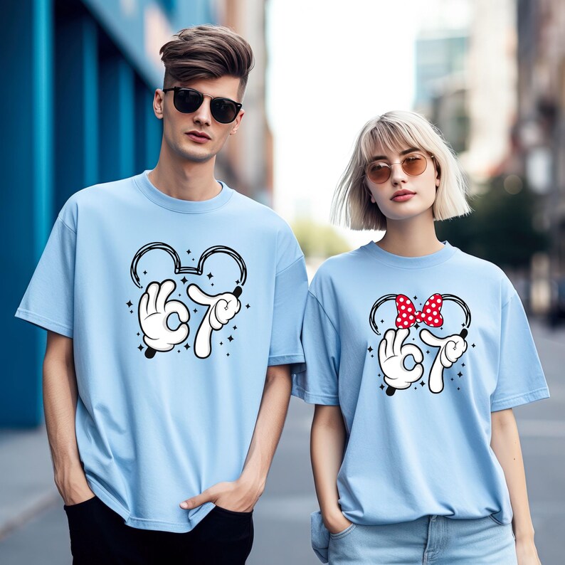 K&ouml;nnte beinhalten: Zwei hellblaue T-Shirts mit einem von Mickey Mouse inspirierten Design. Das Design umfasst eine Mickey Mouse-Kopfkontur, wei&szlig;e behandschuhte H&auml;nde, die die Zahl 67 bilden, und Sternakzente. Ein Shirt hat eine rote Schleife mit Punkten.
