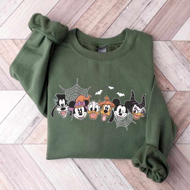 Puede incluir: Una sudadera verde con un estampado de Mickey Mouse y sus amigos disfrazados de Halloween. Los personajes est&aacute;n rodeados de una telara&ntilde;a y murci&eacute;lagos.