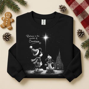 May include: Black sweatshirt featuring a graphic of Mickey Mouse and Pluto looking at a star, with a castle in the background. The text "Believe in the wonder of Christmas" is above the characters. Festive holiday apparel.