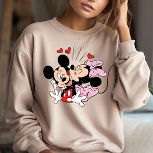 May include: A beige sweatshirt featuring a printed design of Mickey Mouse and Minnie Mouse kissing. The design includes red hearts and a pink bow on Minnie Mouse.
