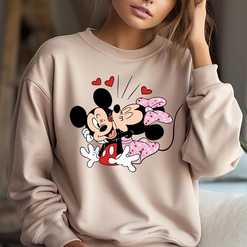 May include: A beige sweatshirt featuring a printed design of Mickey Mouse and Minnie Mouse kissing. The design includes red hearts and a pink bow on Minnie Mouse.