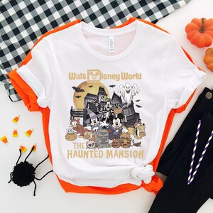 May include: White t-shirt with a graphic of Mickey Mouse and friends in front of the Haunted Mansion at Walt Disney World. The text "Walt Disney World" and "The Haunted Mansion" are printed on the shirt.