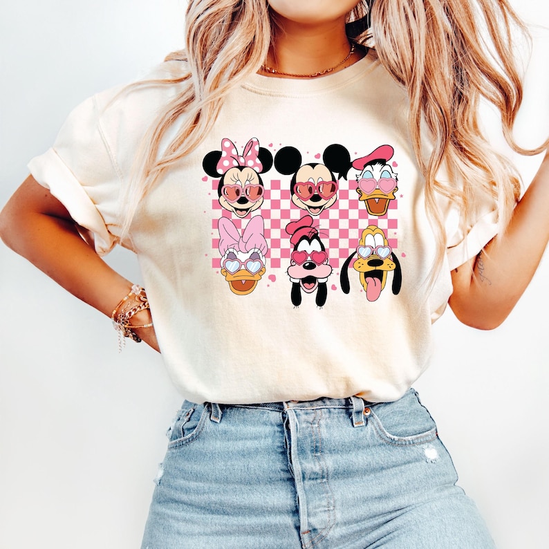 May include: A white t-shirt with a checkered pattern and images of Mickey Mouse, Minnie Mouse, Donald Duck, Goofy, and Pluto wearing heart-shaped sunglasses.