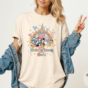 Puede incluir: Camiseta de color marfil con un gr&aacute;fico colorido de personajes de Disney, un arco&iacute;ris y un castillo. El texto "Walt Disney World" est&aacute; impreso debajo. La camiseta tiene mangas cortas y un corte holgado.