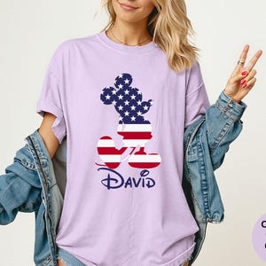 May include: Orchid-colored t-shirt featuring a patriotic Mickey Mouse design. The Mickey Mouse silhouette is filled with a pattern of stars and stripes, with the name "David" in a navy blue script below. The shirt is a Comfort Colors brand.
