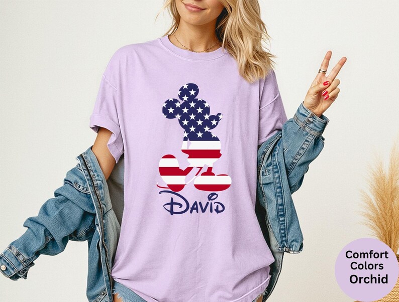 May include: Orchid-colored t-shirt featuring a patriotic Mickey Mouse design. The Mickey Mouse silhouette is filled with a pattern of stars and stripes, with the name "David" in a navy blue script below. The shirt is a Comfort Colors brand.