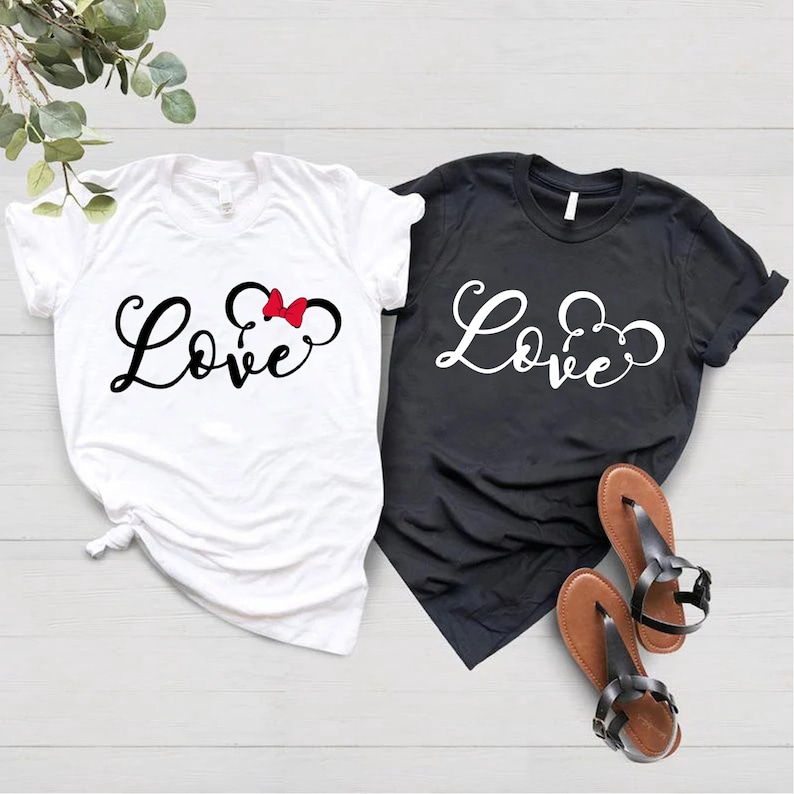 K&ouml;nnte beinhalten: Zwei T-Shirts mit dem Wort "Love" in einer kursiven Schrift. Das wei&szlig;e T-Shirt hat eine rote Schleife und Mickey-Maus-Ohren. Das schwarze T-Shirt hat wei&szlig;e Mickey-Maus-Ohren.