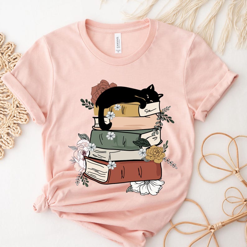 May include: A pink t-shirt with a black cat sleeping on a stack of books. The books are decorated with flowers and leaves.