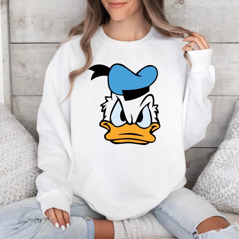 May include: White sweatshirt featuring a cartoon graphic of Donald Duck's face. The design includes a blue hat, black accents, and an orange beak. The sweatshirt is a casual, long-sleeved top.