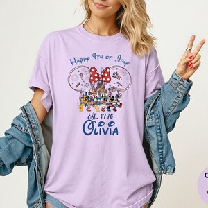 Pode incluir: Uma t-shirt lil&aacute;s clara com um gr&aacute;fico com orelhas do Mickey Mouse, um la&ccedil;o vermelho e o texto "Happy 4th of July Est. 1776 Olivia". O design inclui personagens de desenhos animados e fogos de artif&iacute;cio. A t-shirt faz parte da cole&ccedil;&atilde;o Comfort Colors Orchid.