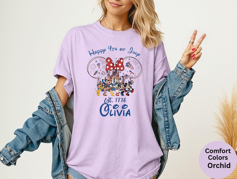 Pode incluir: Uma t-shirt lil&aacute;s clara com um gr&aacute;fico com orelhas do Mickey Mouse, um la&ccedil;o vermelho e o texto "Happy 4th of July Est. 1776 Olivia". O design inclui personagens de desenhos animados e fogos de artif&iacute;cio. A t-shirt faz parte da cole&ccedil;&atilde;o Comfort Colors Orchid.
