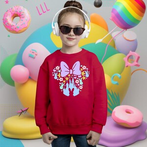 May include: A red sweatshirt featuring a Minnie Mouse-inspired design. The design includes a large purple bow, flowers, bunny silhouettes, and a castle. The sweatshirt is worn by a person wearing sunglasses and headphones. Colorful balloons and donuts are in the background.